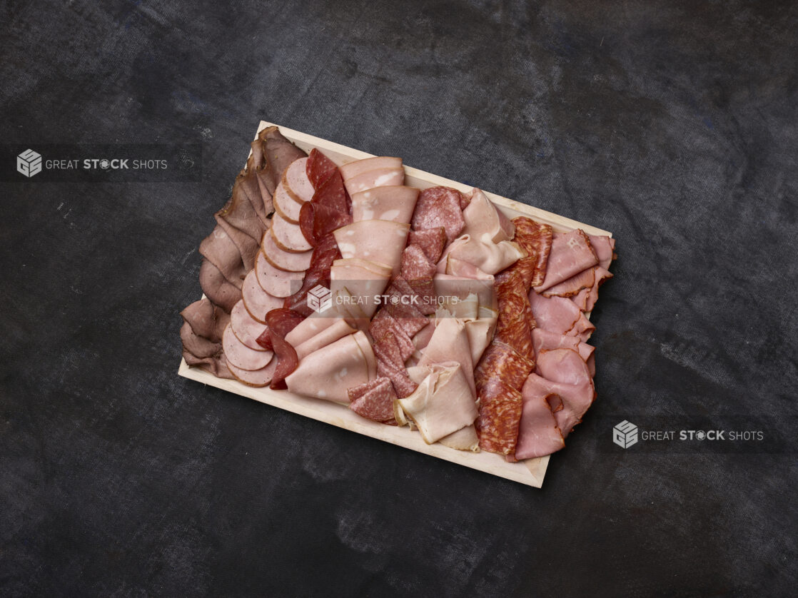 Overhead view of a wooden platter of cured sliced deli meats on a dark background