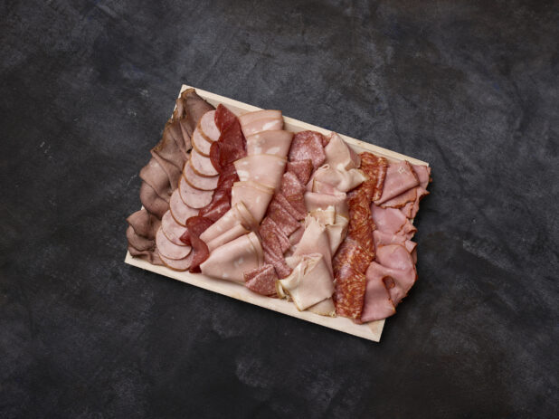 Overhead view of a wooden platter of cured sliced deli meats on a dark background