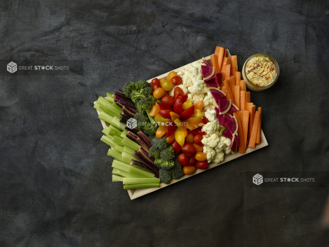 Overhead of raw vegetables for dipping on a wooden tray with a side of hummus in a glass jar on a dark background