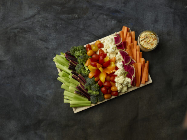 Overhead of raw vegetables for dipping on a wooden tray with a side of hummus in a glass jar on a dark background