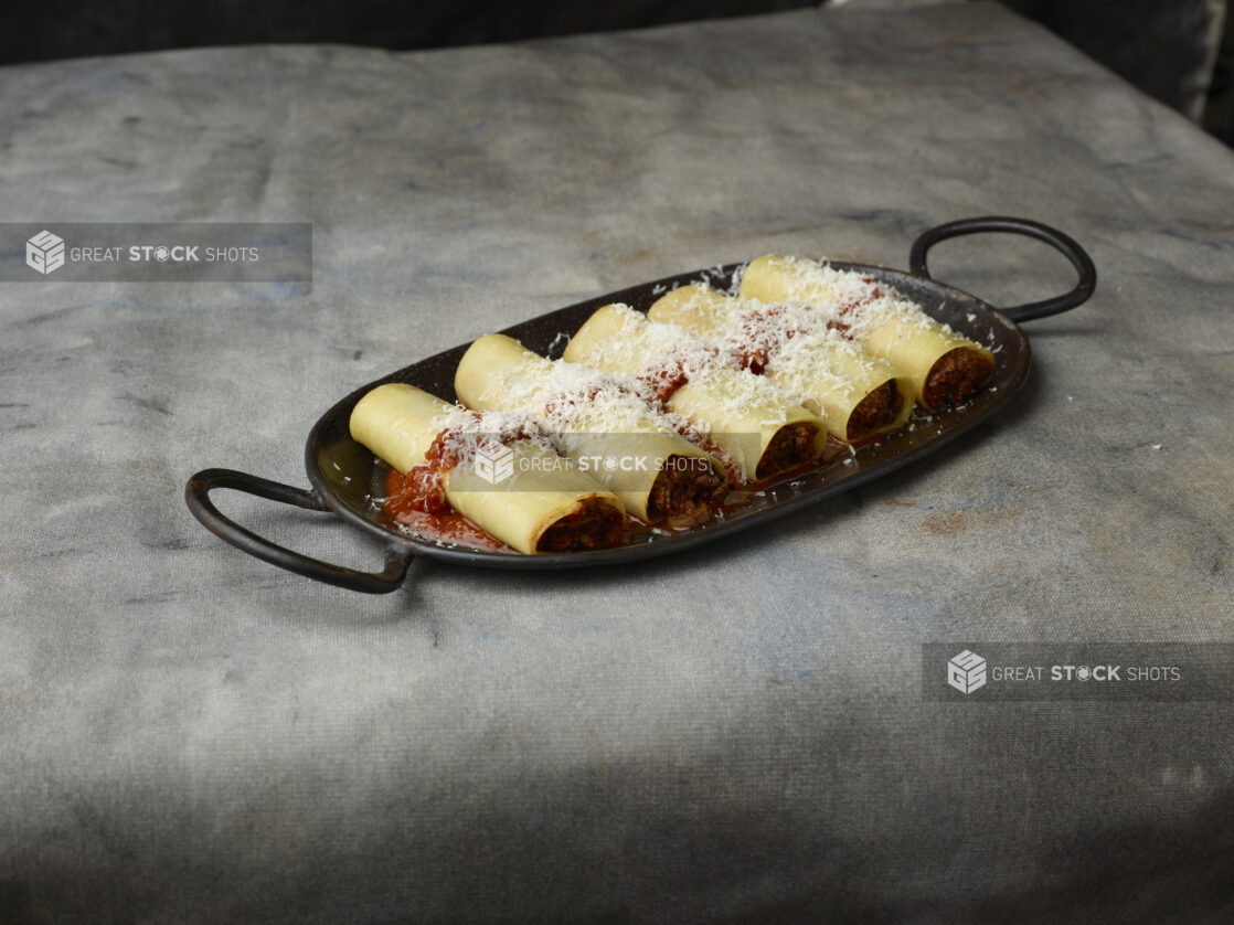 Row of veal cannelloni on a black cast iron tray topped with tomato sauce and freshly grated parmesan cheese