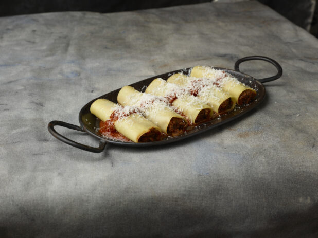 Row of veal cannelloni on a black cast iron tray topped with tomato sauce and freshly grated parmesan cheese