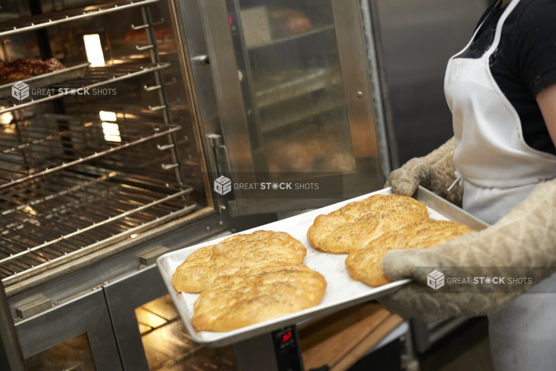 An Employee with Oven Mitts Pulling Out a Baking Sheet of Fresh Baked Mini Herbed Focaccia Bread in the Kitchen of a Gourmet Grocery Store