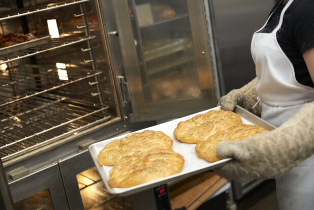 An Employee with Oven Mitts Pulling Out a Baking Sheet of Fresh Baked Mini Herbed Focaccia Bread in the Kitchen of a Gourmet Grocery Store