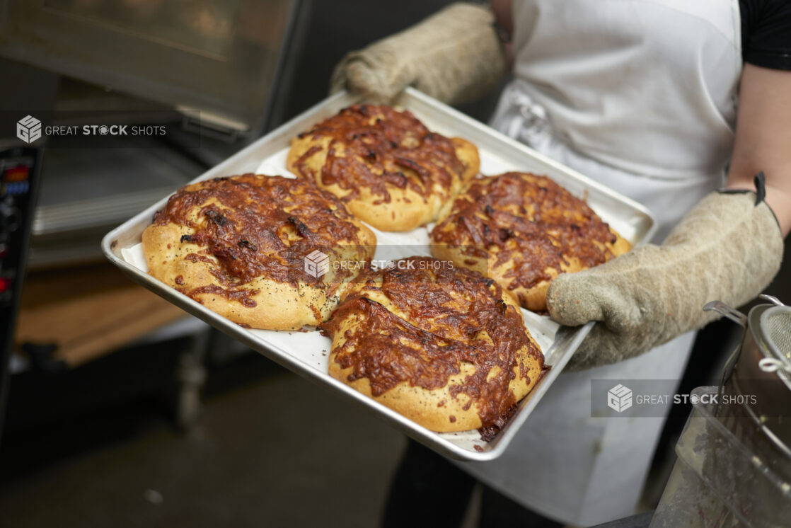 Close Up of an Employee with Oven Mitts Holding a Baking Sheet of Fresh Baked Mini Cheese Buns in the Kitchen of a Gourmet Grocery Store