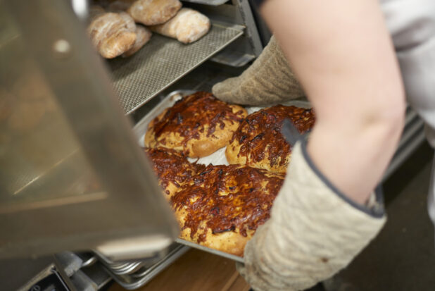 Baker loading a tray of cheesy buns onto a baking rack in a commercial bakery