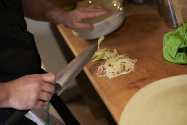 A chef's hand holding a knife over some thinly sliced onions