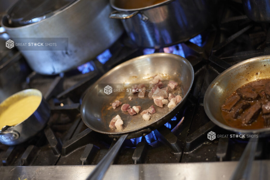 A hot sauté pan with lardons over a blue gas flame