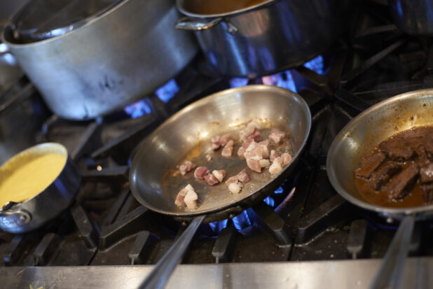 A hot sauté pan with lardons over a blue gas flame