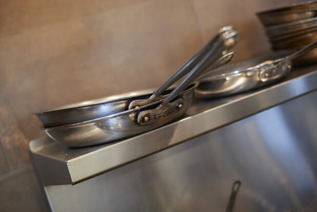 Sautée pans stacked on a steel shelf above a kitchen range, close-up