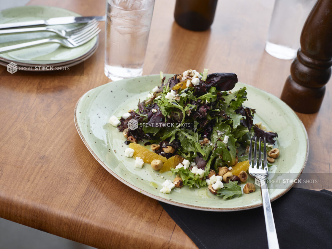 Salad of mixed greens with oranges, hazelnuts, and crumbled white cheese, green plate, wood tabletop