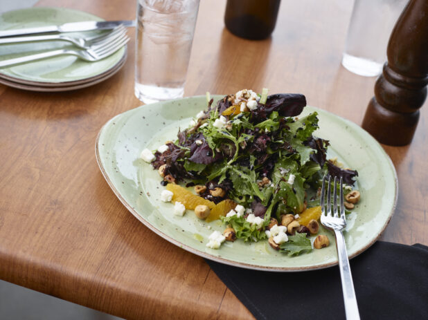 Salad of mixed greens with oranges, hazelnuts, and crumbled white cheese, green plate, wood tabletop