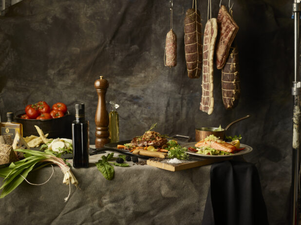 Long table with entrees and quality ingredients with hanging meat in the background with a dark backdrop