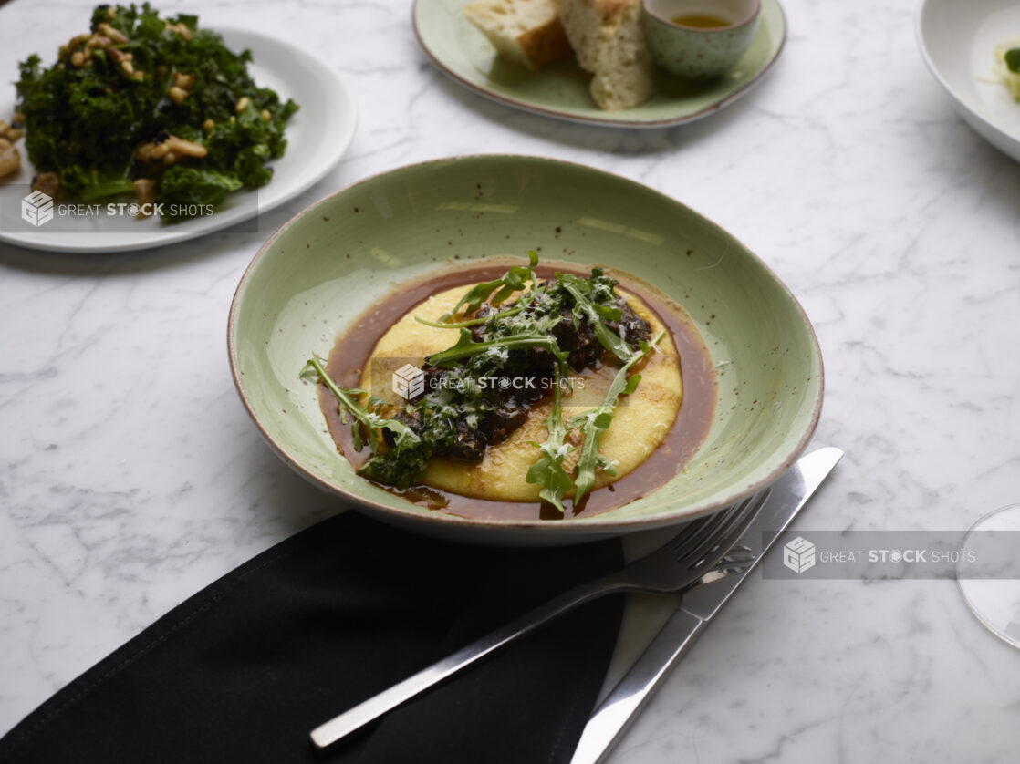 Braised Beef Short Ribs and Baby Arugula on a Bed of Polenta with Au Jus in a Green Ceramic Dish on a Marble Table