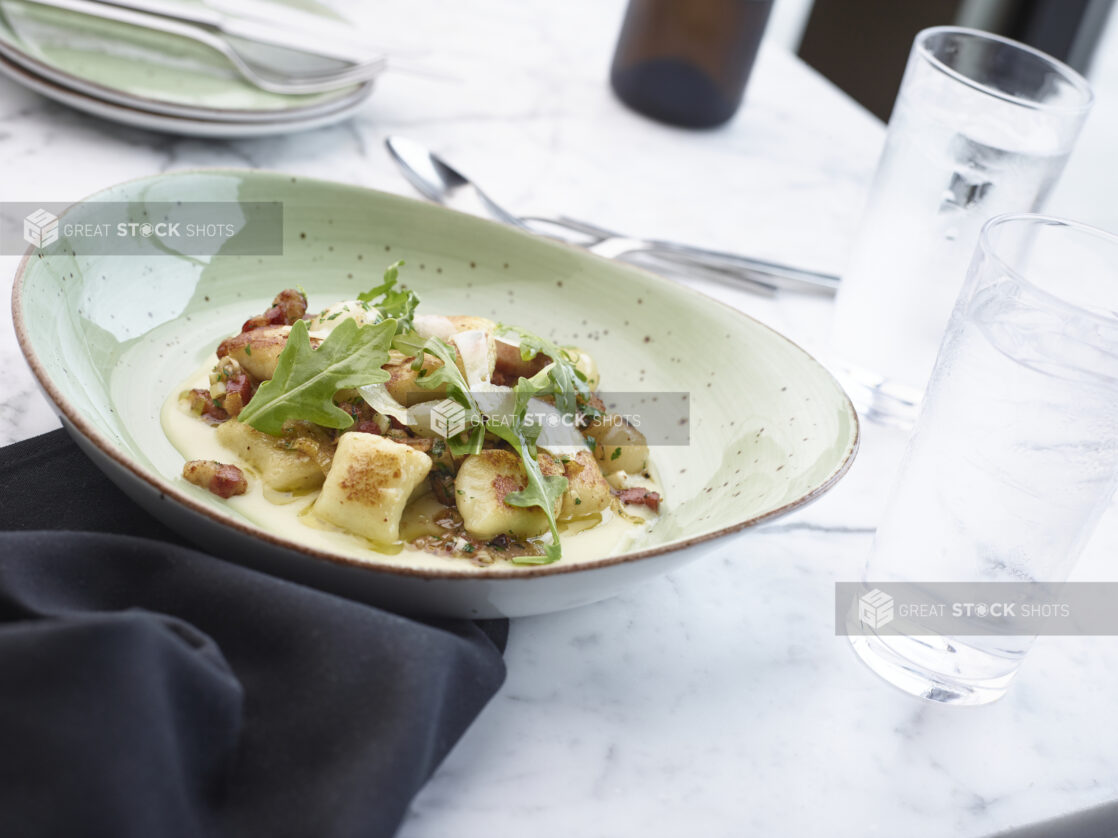 Large green bowl of creamy gnocchi and bacon garnished with arugula and shaved parmesan cheese