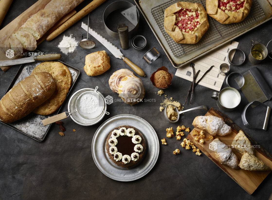 Overhead View of Assorted Breads, Pastries, Cakes, Pies and Baked Goods on a Dark Canvas Surface with Baking Utensils