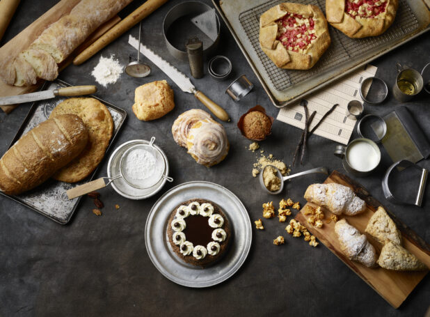Overhead View of Assorted Breads, Pastries, Cakes, Pies and Baked Goods on a Dark Canvas Surface with Baking Utensils
