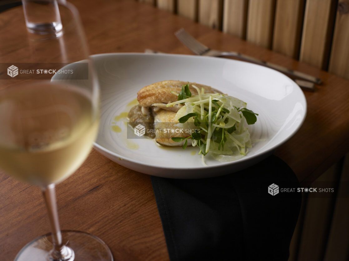 Pan-fried white fish with fennel and microgreens garnish, glass of white wine in foreground