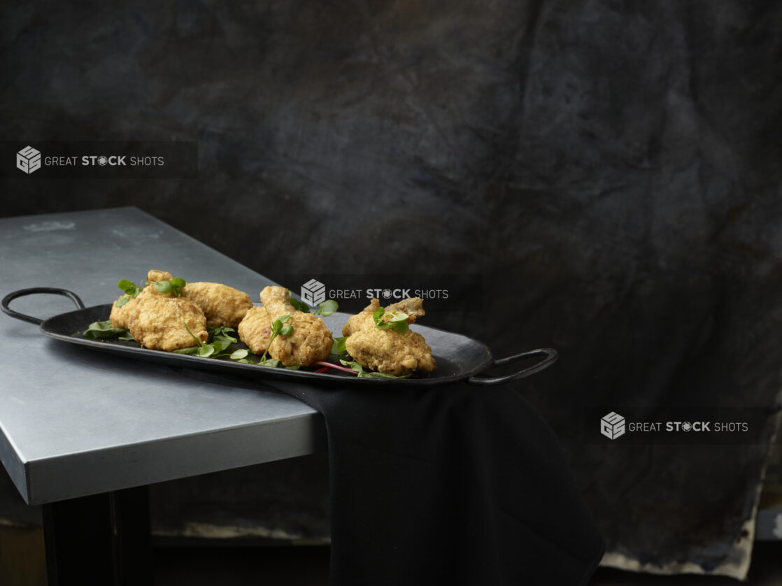 Black serving platter of fried tuscan chicken garnished with greens on a metal table and a black napkin