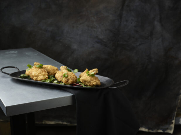Black serving platter of fried tuscan chicken garnished with greens on a metal table and a black napkin