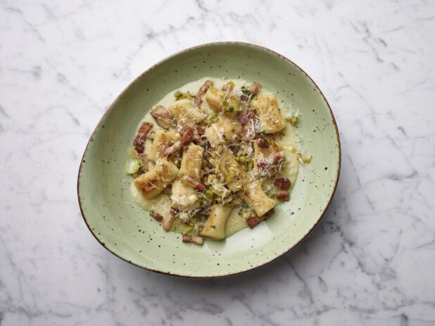 Overhead bowl of gnocchi with pancetta and shaved parmesan in a cream sauce on a white marble background