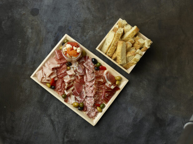 Overhead of sliced cured meats on a wooden tray with a side of focaccia on a dark background