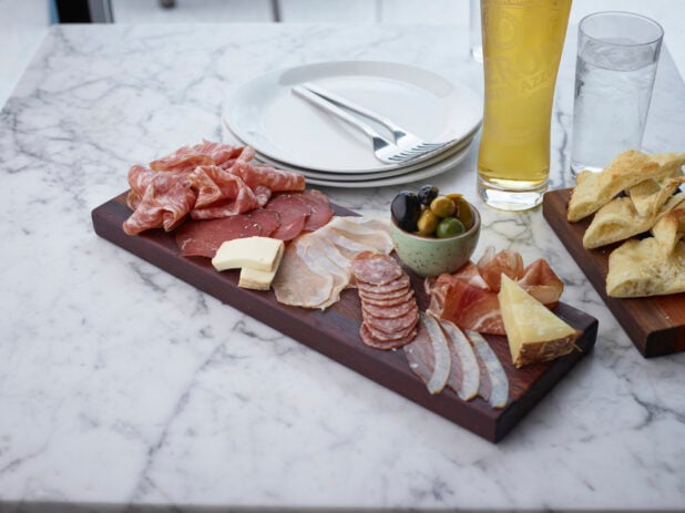 Charcuterie board with Italian cured meats, cheeses, and olives, pint of lager in background on a white marble tabletop