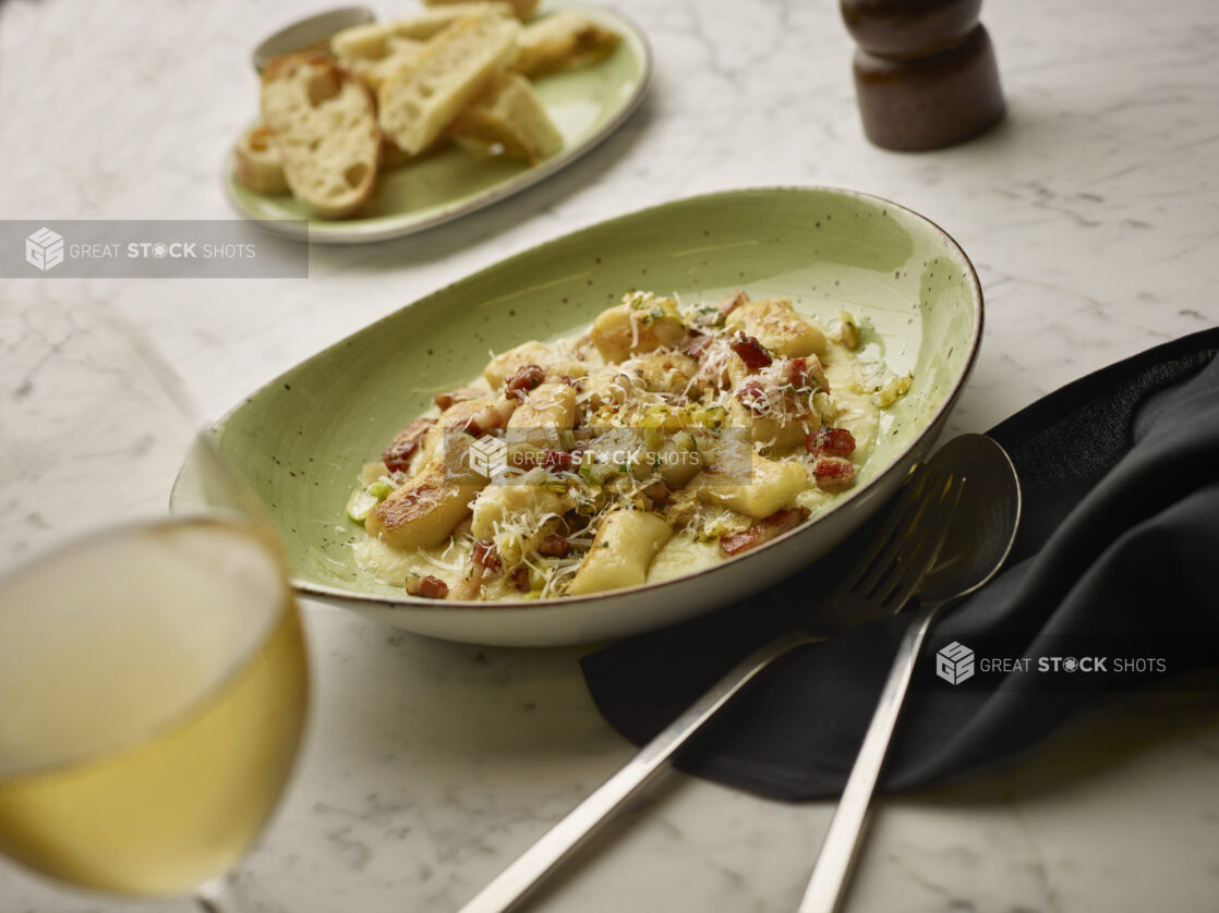 Bowl of gnocchi with pancetta and shaved parmesan in a cream sauce with a plate of bread in the background