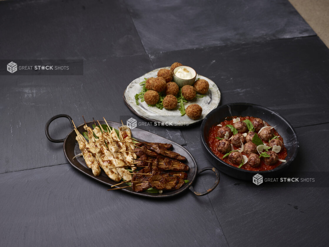 Collection of appetizers, chicken and beef satay, meatballs and arancini on rustic serving dishes on a dark background