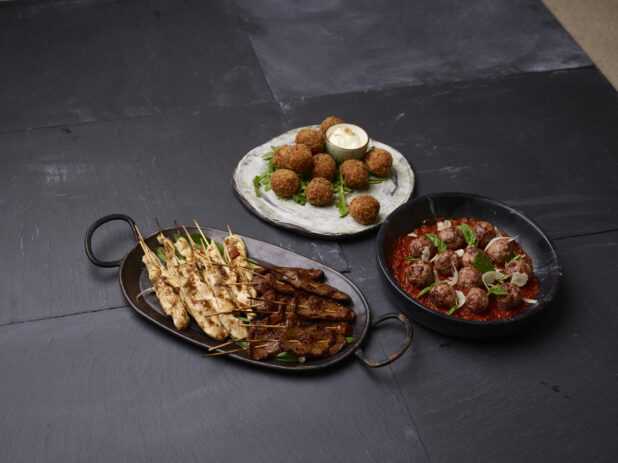 Collection of appetizers, chicken and beef satay, meatballs and arancini on rustic serving dishes on a dark background