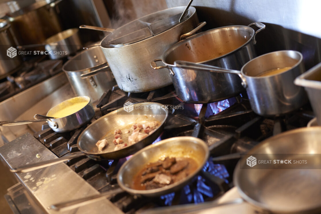 Sauté pans and stock pots on a gas range with blue flames in a commercial kitchen