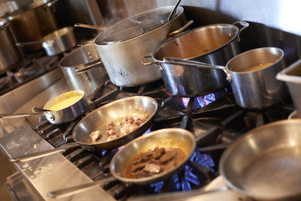 Sauté pans and stock pots on a gas range with blue flames in a commercial kitchen