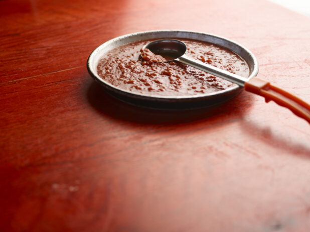 Metal pan of pizza sauce on a red wooden table with ladle scooping sauce