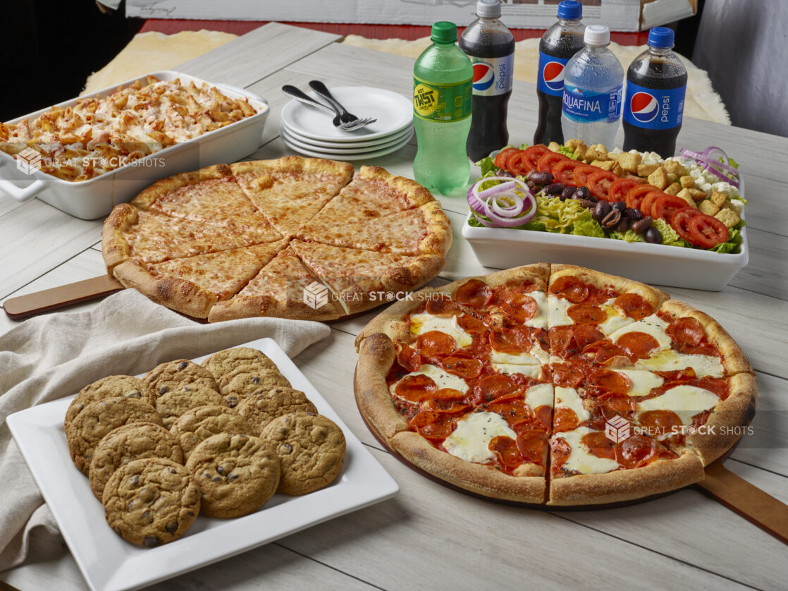 Family style meal with two pizzas, greek salad, baked penne with cheese, dozen cookies and drinks on a wooden background