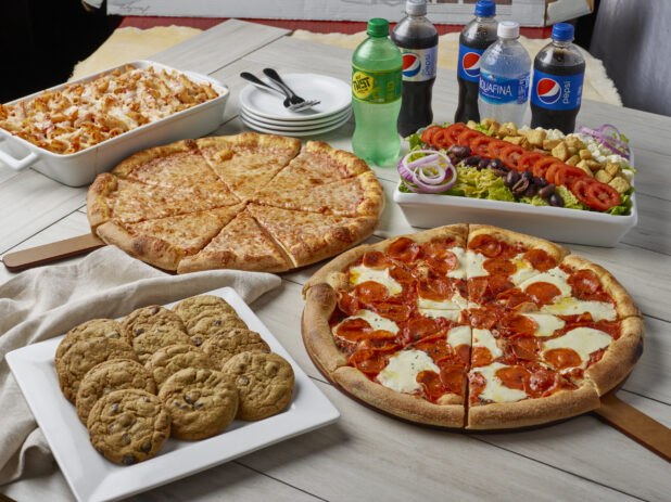 Family style meal with two pizzas, greek salad, baked penne with cheese, dozen cookies and drinks on a wooden background