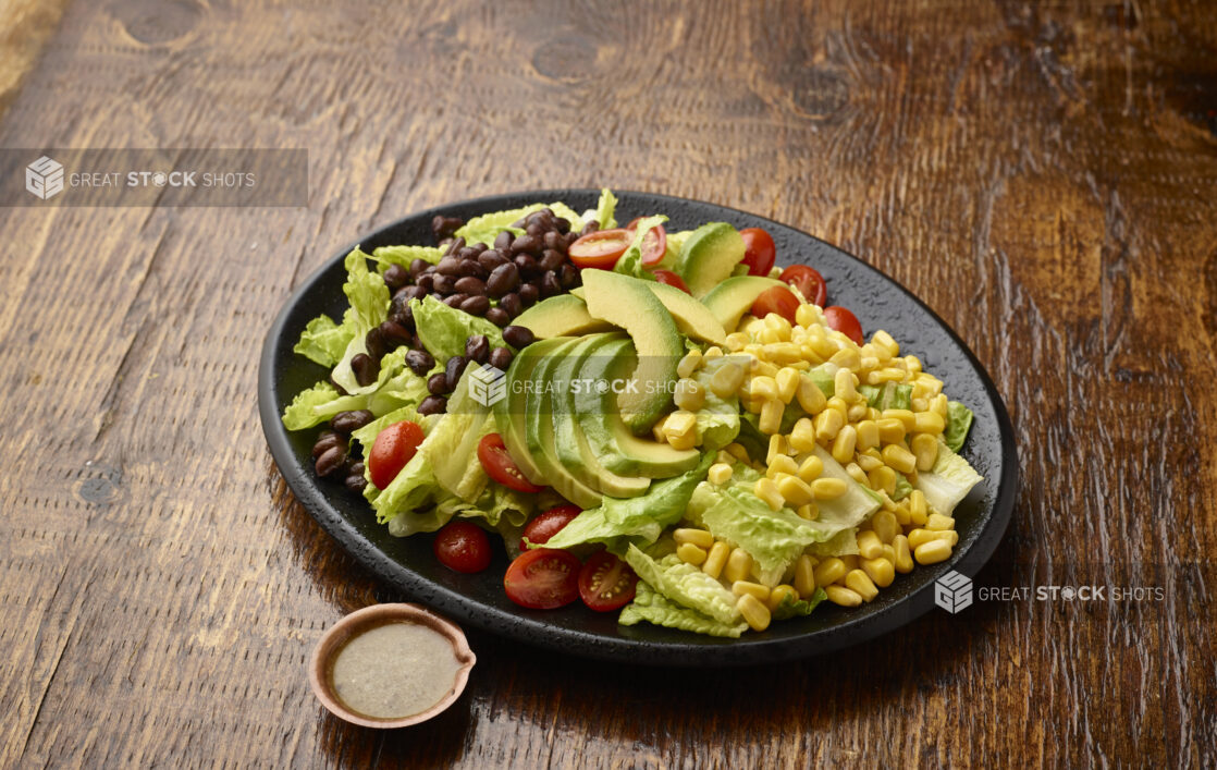 Plain Tex-Mex Salad of Black Beans, Avocados and Corn with a Side of Salad Dressing on a Black Ceramic Plate on a Dark Wood Table