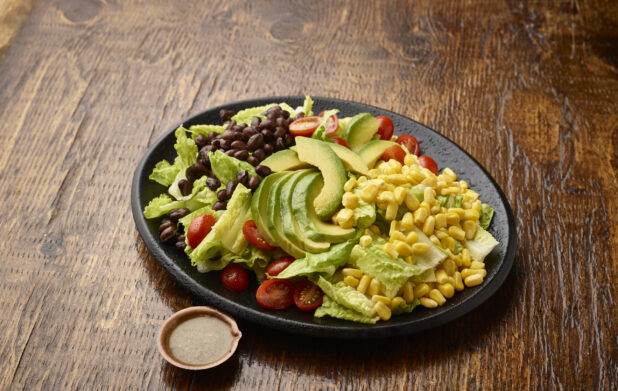 Plain Tex-Mex Salad of Black Beans, Avocados and Corn with a Side of Salad Dressing on a Black Ceramic Plate on a Dark Wood Table