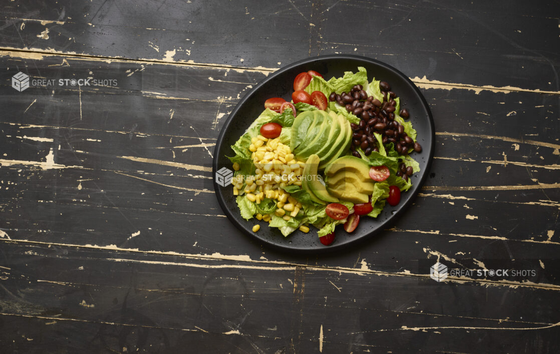 Overhead View of a Tex Mex Style Garden Salad with Avocado Slices, Black Beans and Corn on a Black Ceramic Platter on a Black Painted Wood Table