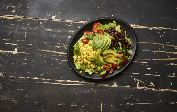 Overhead View of a Tex Mex Style Garden Salad with Avocado Slices, Black Beans and Corn on a Black Ceramic Platter on a Black Painted Wood Table