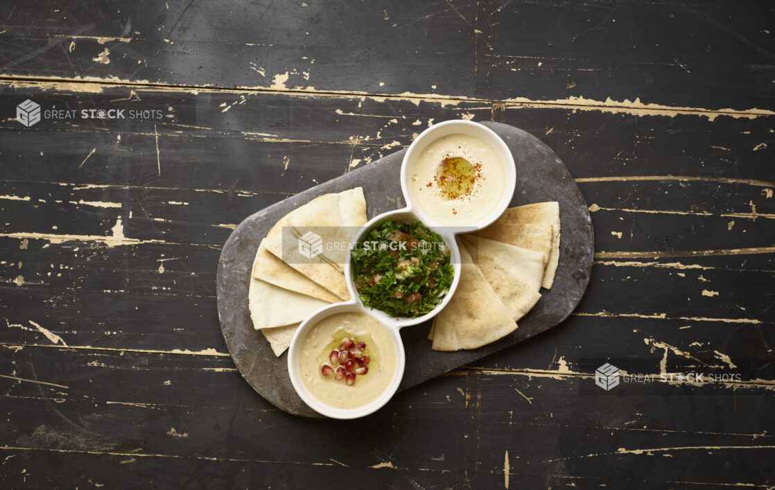 Overhead View of a Trio Plate of Side Dishes: Tabbouleh, Baba Ganoush and Hummus with Pita Bread Wedges and a Black Slate Platter on a Black Painted Wood Table