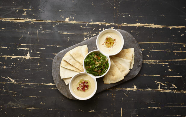 Overhead View of a Trio Plate of Side Dishes: Tabbouleh, Baba Ganoush and Hummus with Pita Bread Wedges and a Black Slate Platter on a Black Painted Wood Table