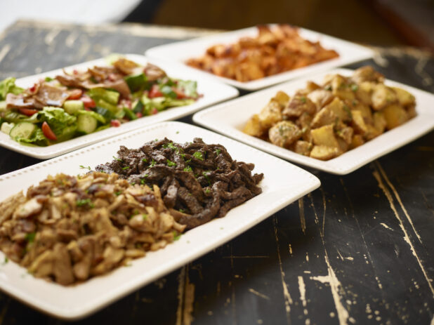 A Family Dining or Catering Spread of Middle Eastern Foods: Beef and Chicken Shawarma, Chicken Kebabs, Garlic Potatoes and a Gyro Salad in White Ceramic Platters on a Black Painted Wood Table