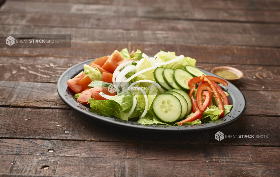 Plain Garden Salad with a Side of Salad Dressing on a Black Ceramic Plate on a Dark Wood Panelled Table