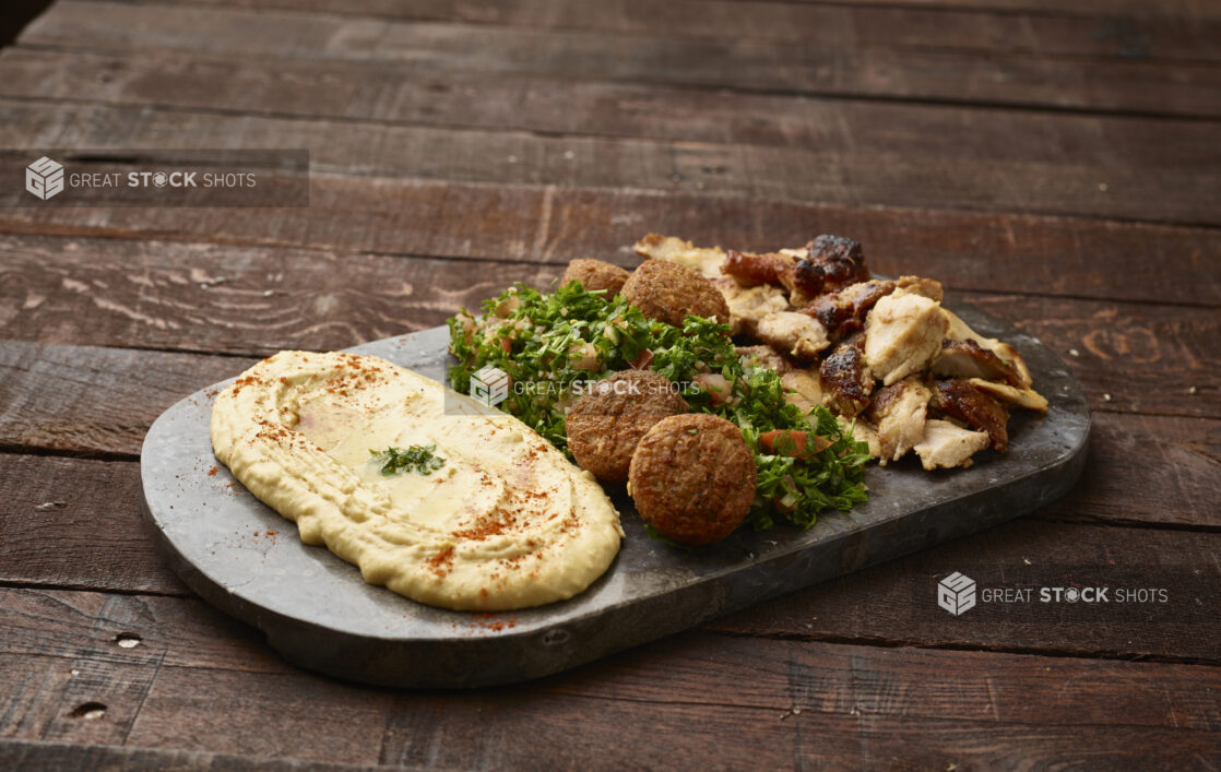 Sharing Plate: Tabbouleh, Hummus, Falafel Balls and Chicken Shawarma on a Black Slate Platter on a Dark Wood Panelled Table