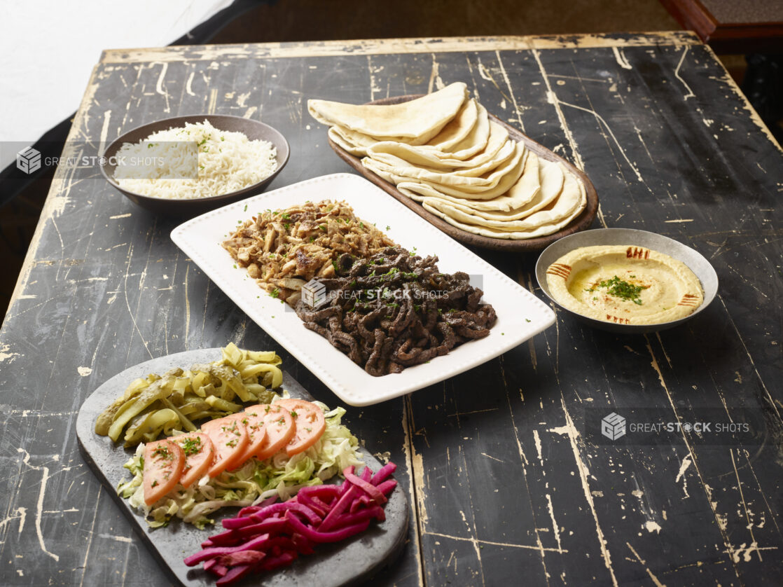 Build Your Own Pita Wrap Set with Beef and Chicken Shawarma, Vegetable Toppings, Pita Bread and Sides of White Rice and Hummos Dip on a Black Painted Wooden Table