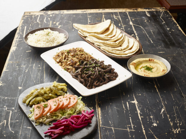 Build Your Own Pita Wrap Set with Beef and Chicken Shawarma, Vegetable Toppings, Pita Bread and Sides of White Rice and Hummos Dip on a Black Painted Wooden Table