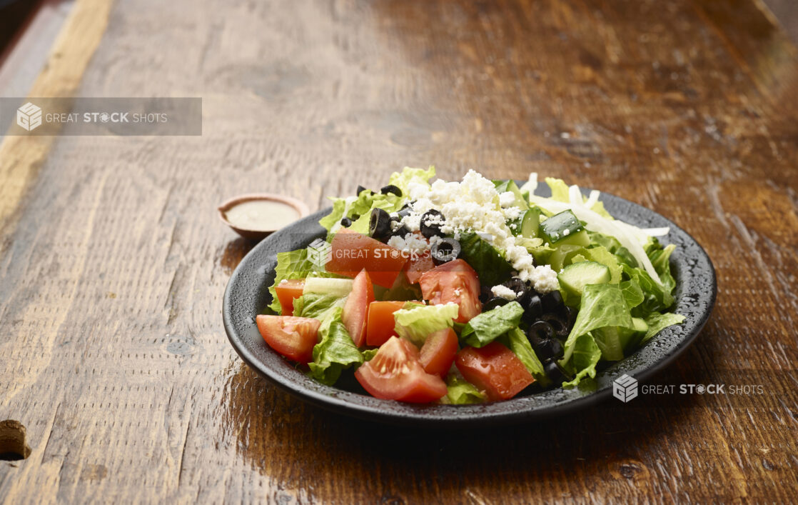 Plain Greek Salad with a Side of Salad Dressing on a Black Ceramic Plate on a Dark Wood Table