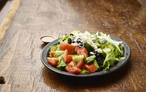 Plain Greek Salad with a Side of Salad Dressing on a Black Ceramic Plate on a Dark Wood Table