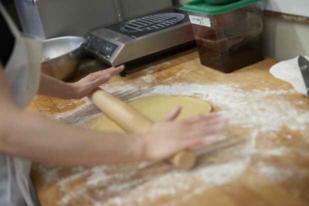 Baker rolling out dough on a floured wood board in a commercial bakery