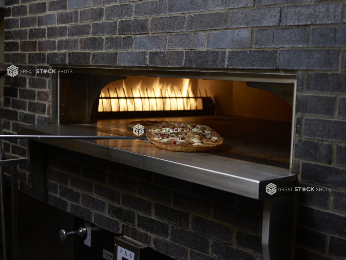 Whole Pizza on a Pizza Peel Coming Out of a Wood Fired Pizza Oven in a Restaurant Kitchen Setting